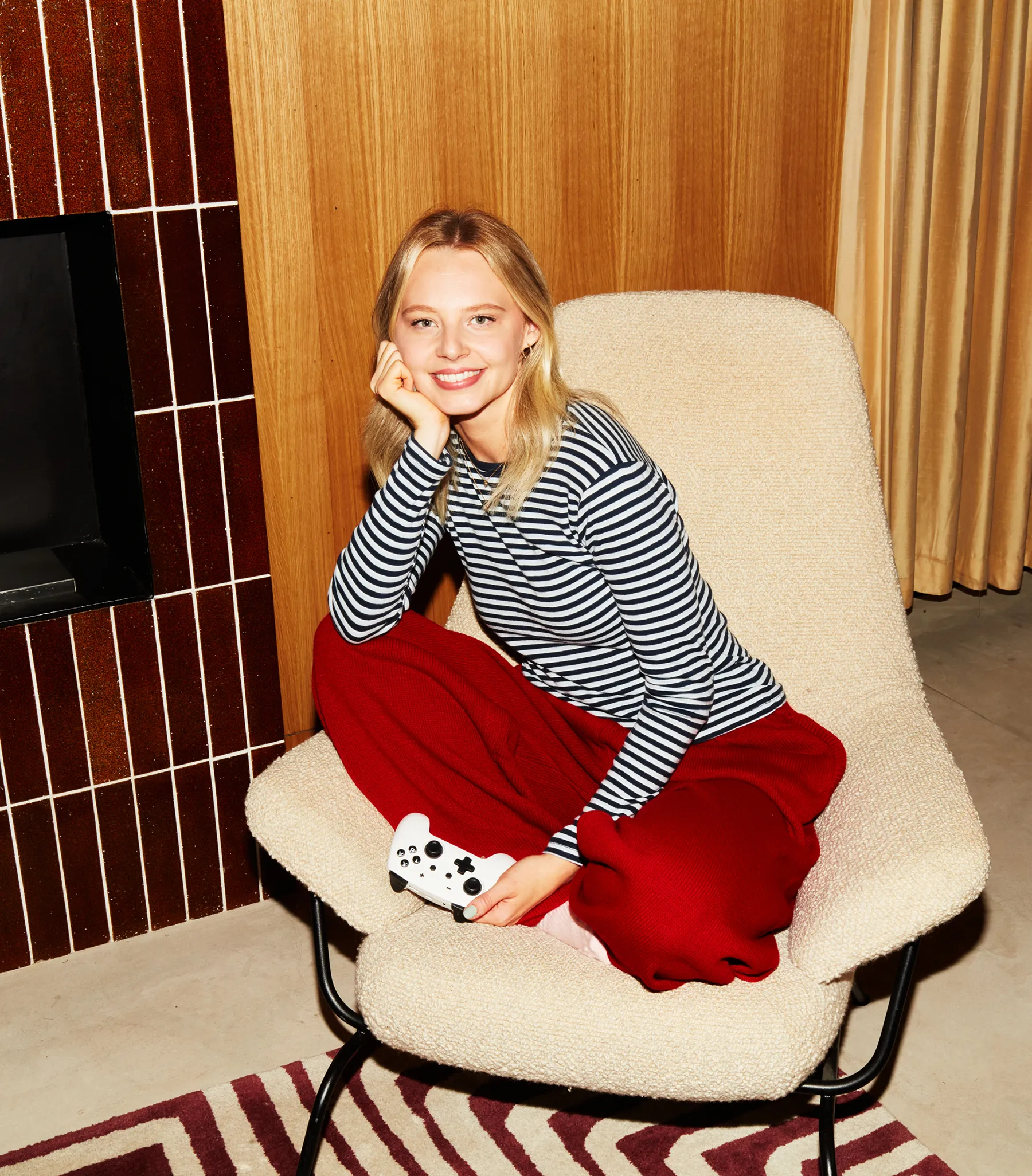 Young woman in striped top and red pants sitting in cream chair holding white gaming controller, smiling at camera in modern living room setting.